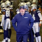 FootballR - NFL - Im Vordergrund steht Mike McCarthy in marineblauer Jacke und Mütze mit dem Mannschaftslogo der Dallas Cowboys. Hinter ihm versammeln sich Spieler in blau-weißen Uniformen mit Helmen in einem Tunnel und sind bereit, das Stadion zu betreten. Links ist ein Mitarbeiter in einer blauen Weste zu sehen. Diese Beschreibung wurde automatisch generiert.