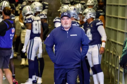 FootballR - NFL - Im Vordergrund steht Mike McCarthy in marineblauer Jacke und Mütze mit dem Mannschaftslogo der Dallas Cowboys. Hinter ihm versammeln sich Spieler in blau-weißen Uniformen mit Helmen in einem Tunnel und sind bereit, das Stadion zu betreten. Links ist ein Mitarbeiter in einer blauen Weste zu sehen. Diese Beschreibung wurde automatisch generiert.