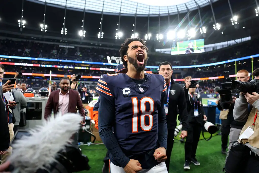 Black Friday - NFL - Caleb Williams, der ein dunkles Trikot mit der Nummer 18 trägt, jubelt ausgelassen auf dem Feld, umgeben von Fotografen und Journalisten. Das Stadion ist hell erleuchtet, im Hintergrund ist eine große Leinwand zu sehen, auf den Tribünen sitzen Zuschauer. Sein Gesichtsausdruck ist geprägt von Triumph und Aufregung. Diese Beschreibung wurde automatisch generiert.