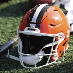 FootballR - NFL- Denzel Ward - Ein orangefarbener Footballhelm der Cleveland Browns mit einem breiten weißen Streifen, flankiert von zwei schwarzen Streifen, liegt auf einem Kunstrasenplatz in der Mega-Arena. Neben dem Helm liegen ein schwarzer Gummihammer und ein Paar weiße Footballhandschuhe. Diese Beschreibung wurde automatisch generiert.