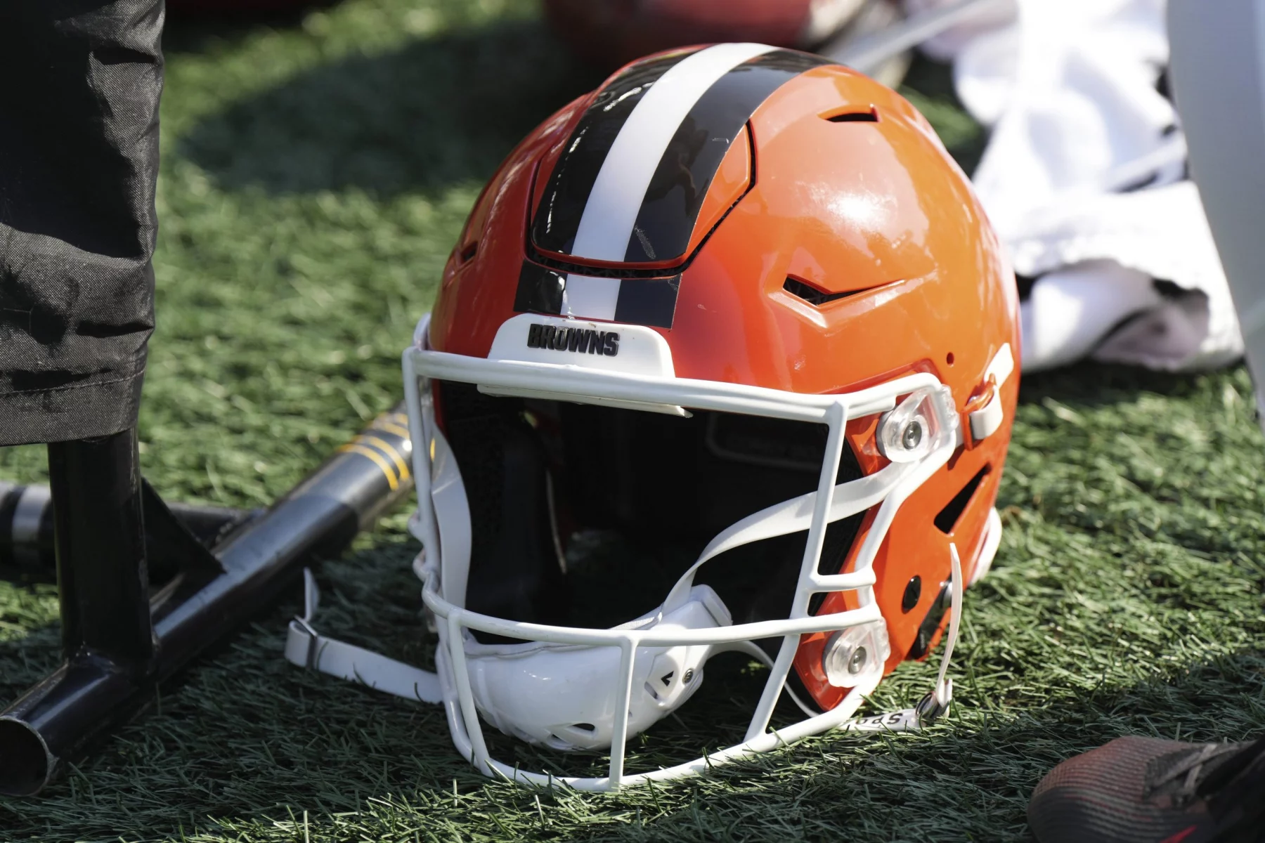 FootballR - NFL- Denzel Ward - Ein orangefarbener Footballhelm der Cleveland Browns mit einem breiten weißen Streifen, flankiert von zwei schwarzen Streifen, liegt auf einem Kunstrasenplatz in der Mega-Arena. Neben dem Helm liegen ein schwarzer Gummihammer und ein Paar weiße Footballhandschuhe. Diese Beschreibung wurde automatisch generiert.