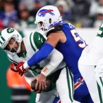 FootballR - NFL - Aaron Rodgers von den New York Jets in grün-weißer Uniform wird von einem Spieler der Buffalo Bills in blauer Uniform mit weißem Helm mit roten Details angegriffen. Die Aktion findet während eines AFC East-Spiels statt, wobei andere Spieler in ähnlichen Trikots teilweise im Hintergrund zu sehen sind. Diese Beschreibung wurde automatisch generiert.