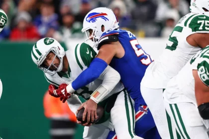 FootballR - NFL - Aaron Rodgers von den New York Jets in grün-weißer Uniform wird von einem Spieler der Buffalo Bills in blauer Uniform mit weißem Helm mit roten Details angegriffen. Die Aktion findet während eines AFC East-Spiels statt, wobei andere Spieler in ähnlichen Trikots teilweise im Hintergrund zu sehen sind. Diese Beschreibung wurde automatisch generiert.