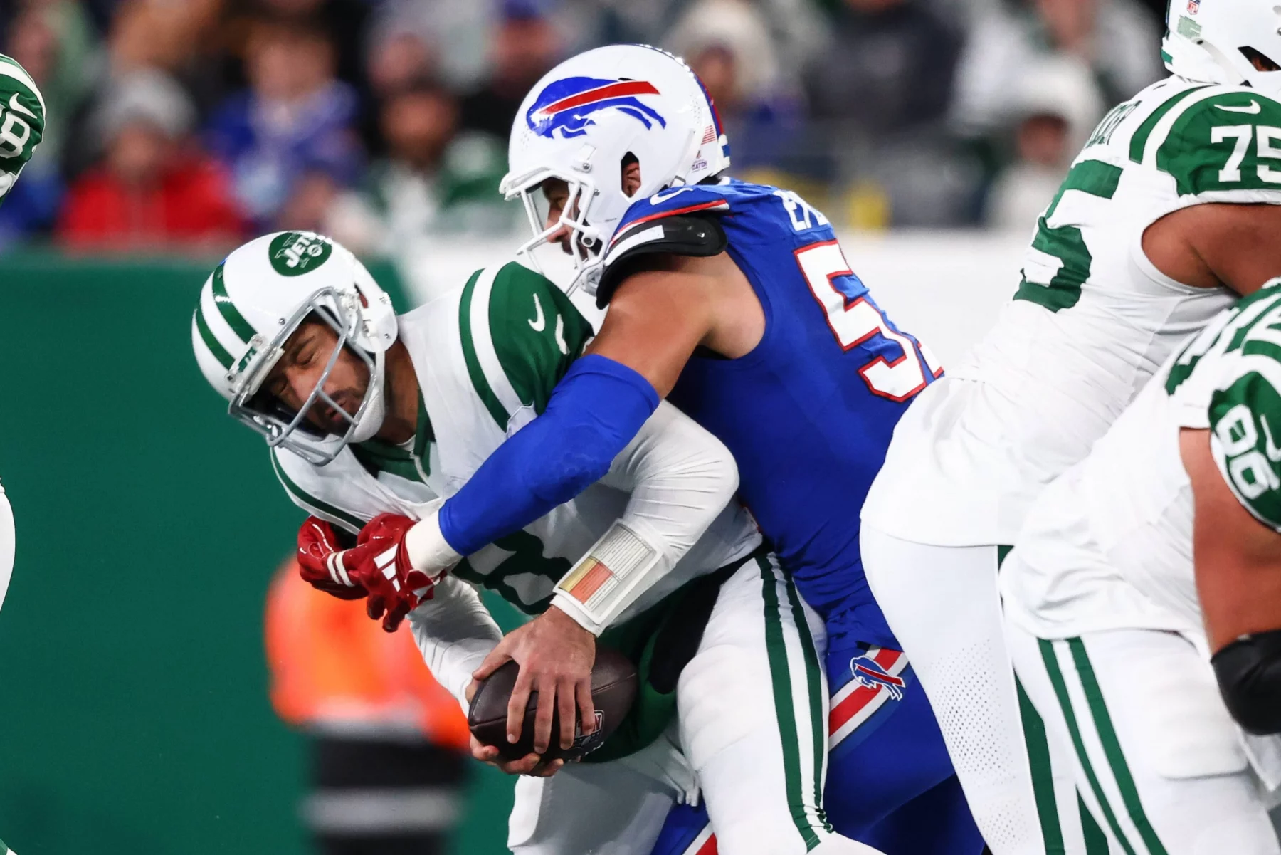 FootballR - NFL - Aaron Rodgers von den New York Jets in grün-weißer Uniform wird von einem Spieler der Buffalo Bills in blauer Uniform mit weißem Helm mit roten Details angegriffen. Die Aktion findet während eines AFC East-Spiels statt, wobei andere Spieler in ähnlichen Trikots teilweise im Hintergrund zu sehen sind. Diese Beschreibung wurde automatisch generiert.