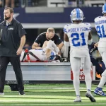 FootballR - NFL - Aidan Hutchinson, der möglicherweise an einem Beinbruch leidet, wird auf einem Wagen medizinisch versorgt. Zwei Teamkollegen in weiß-blauen Lions-Trikots, Nummer 21 und 91, gehen vorbei. Trainer und Mitarbeiter haben sich um das Stadion versammelt, im Hintergrund ein Stadion voller Zuschauer, die auf die Rückkehr ihres Lions-Stars hoffen. Diese Beschreibung wurde automatisch generiert.