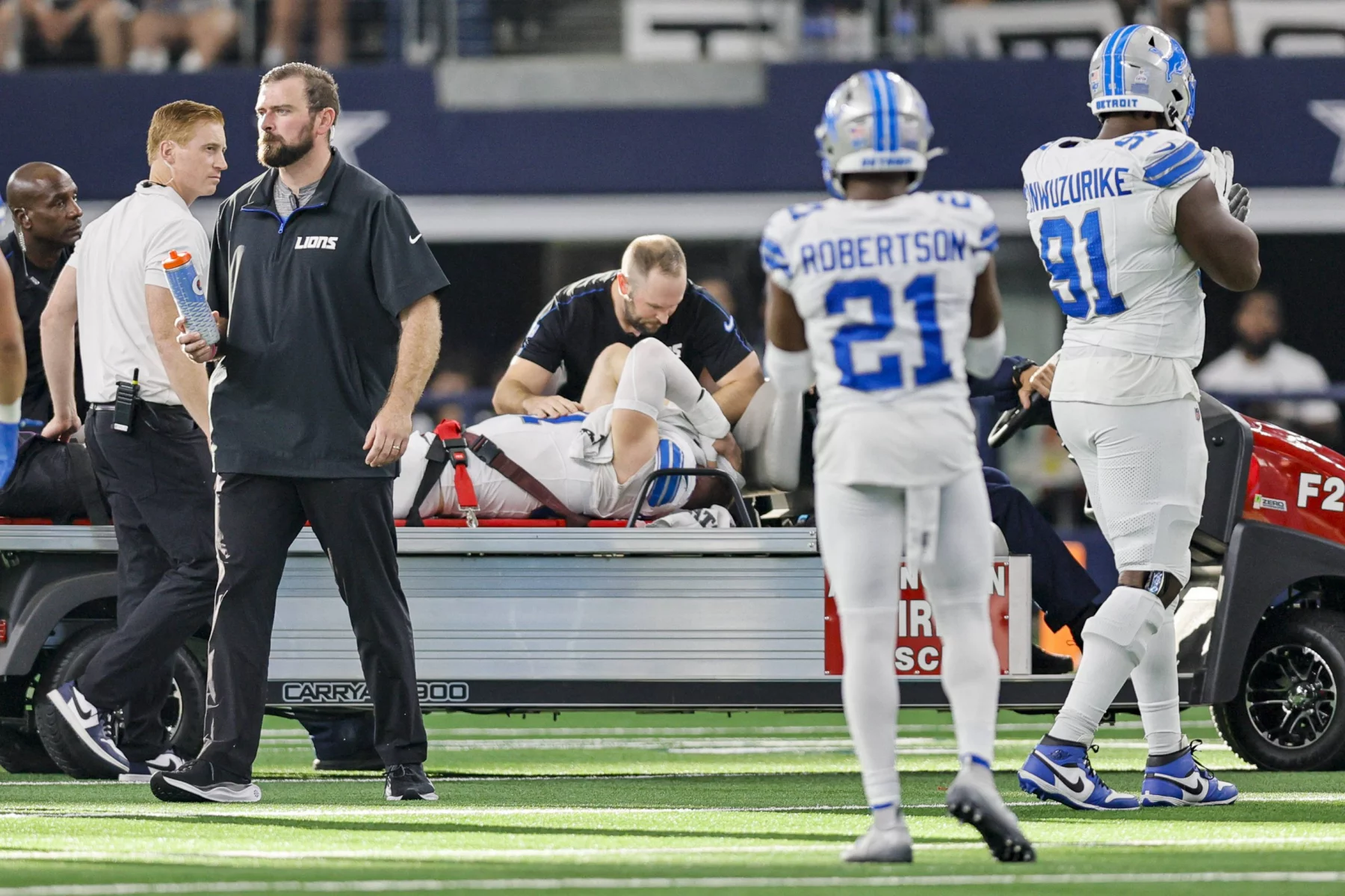 FootballR - NFL - Aidan Hutchinson, der möglicherweise an einem Beinbruch leidet, wird auf einem Wagen medizinisch versorgt. Zwei Teamkollegen in weiß-blauen Lions-Trikots, Nummer 21 und 91, gehen vorbei. Trainer und Mitarbeiter haben sich um das Stadion versammelt, im Hintergrund ein Stadion voller Zuschauer, die auf die Rückkehr ihres Lions-Stars hoffen. Diese Beschreibung wurde automatisch generiert.
