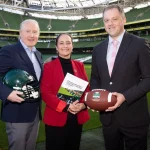 FootballR - NFL International Series Irland - Drei Personen stehen auf einem Footballfeld in einem großen Stadion. Der Mann links hält einen grünen American-Football-Helm und deutet damit auf die NFL-Erweiterung hin. Die Frau in der Mitte scheint mit ihrem Ordner Pläne für International Series-Spiele in Irland zu besprechen, während der Mann rechts einen Football hält. Das Stadion ist leer und das hellgrüne Feld und die Sitze sind sichtbar. Diese Beschreibung wurde automatisch generiert.