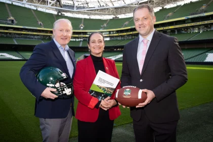 FootballR - NFL International Series Irland - Drei Personen stehen auf einem Footballfeld in einem großen Stadion. Der Mann links hält einen grünen American-Football-Helm und deutet damit auf die NFL-Erweiterung hin. Die Frau in der Mitte scheint mit ihrem Ordner Pläne für International Series-Spiele in Irland zu besprechen, während der Mann rechts einen Football hält. Das Stadion ist leer und das hellgrüne Feld und die Sitze sind sichtbar. Diese Beschreibung wurde automatisch generiert.
