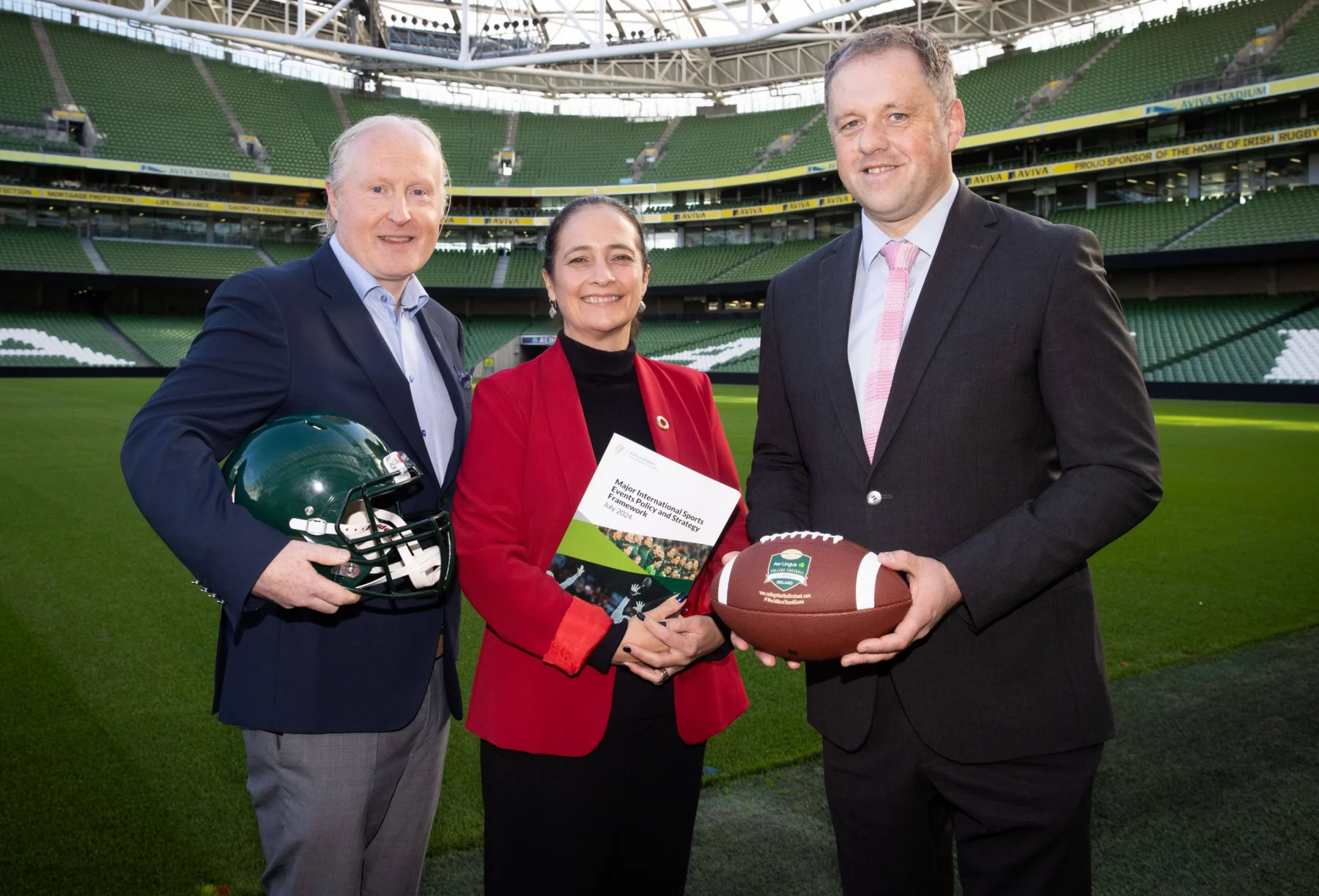 FootballR - NFL International Series Irland - Drei Personen stehen auf einem Footballfeld in einem großen Stadion. Der Mann links hält einen grünen American-Football-Helm und deutet damit auf die NFL-Erweiterung hin. Die Frau in der Mitte scheint mit ihrem Ordner Pläne für International Series-Spiele in Irland zu besprechen, während der Mann rechts einen Football hält. Das Stadion ist leer und das hellgrüne Feld und die Sitze sind sichtbar. Diese Beschreibung wurde automatisch generiert.