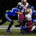 FootballR - NFL - Ein Footballspieler in einer rot-goldenen Uniform, die an einen Athleten der Florida State erinnert, wird von drei Duke-Spielern in Blau und Weiß angegriffen. Der Ball liegt bei schwacher Beleuchtung frei auf dem grünen Feld und bereitet die Bühne für einen historischen Triumph. Diese Beschreibung wurde automatisch generiert.