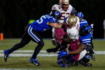 FootballR - NFL - Ein Footballspieler in einer rot-goldenen Uniform, die an einen Athleten der Florida State erinnert, wird von drei Duke-Spielern in Blau und Weiß angegriffen. Der Ball liegt bei schwacher Beleuchtung frei auf dem grünen Feld und bereitet die Bühne für einen historischen Triumph. Diese Beschreibung wurde automatisch generiert.