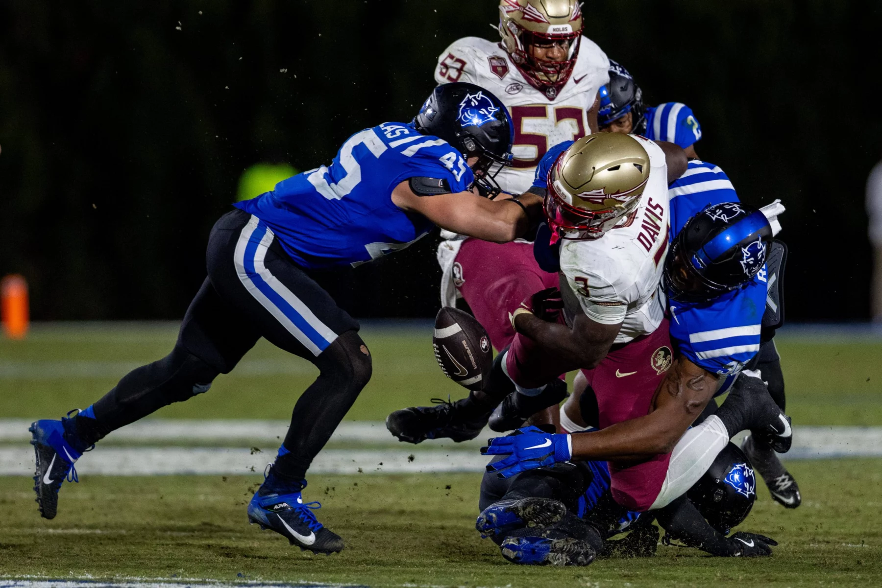 FootballR - NFL - Ein Footballspieler in einer rot-goldenen Uniform, die an einen Athleten der Florida State erinnert, wird von drei Duke-Spielern in Blau und Weiß angegriffen. Der Ball liegt bei schwacher Beleuchtung frei auf dem grünen Feld und bereitet die Bühne für einen historischen Triumph. Diese Beschreibung wurde automatisch generiert.