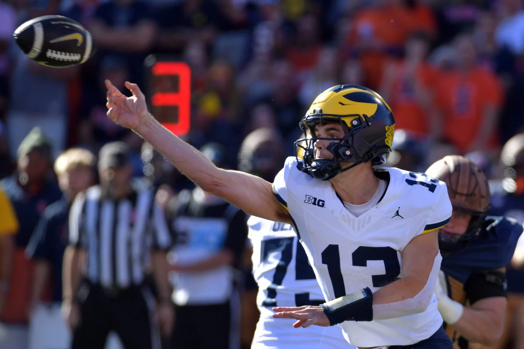 Ein Footballspieler, Jack Tuttle, trägt eine blau-gelbe Uniform mit der Nummer 13 und wirft während eines Spiels einen Football. Er wurde mitten im Wurf mit konzentriertem Gesichtsausdruck aufgenommen, während er spielt, während die digitale Anzeigetafel die Nummer 3 anzeigt. Andere Spieler und ein Schiedsrichter verschwimmen im Hintergrund. Diese Beschreibung wurde mit der FootballR KI automatisch generiert.
