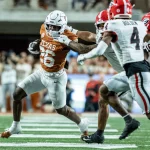 FootballR - NFL - Ein texanischer Footballspieler in Weiß und dunklem Orange versucht während des Spiels, einen Georgia Bulldog in Rot, Schwarz und Weiß mit ausgestrecktem Arm zu überwältigen. Auf dem Rasenfeld, umgeben von verschwommenen Zuschauern, hält der Austin-Athlet den Football in seinem rechten Arm und drängt sich durch die gegnerischen Spieler. Diese Beschreibung wurde automatisch generiert.