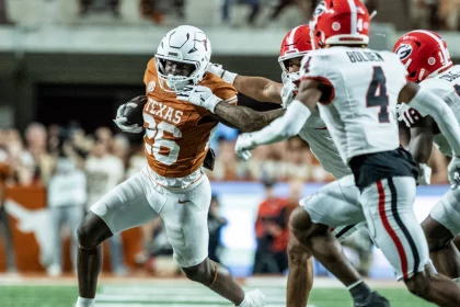 FootballR - NFL - Ein texanischer Footballspieler in Weiß und dunklem Orange versucht während des Spiels, einen Georgia Bulldog in Rot, Schwarz und Weiß mit ausgestrecktem Arm zu überwältigen. Auf dem Rasenfeld, umgeben von verschwommenen Zuschauern, hält der Austin-Athlet den Football in seinem rechten Arm und drängt sich durch die gegnerischen Spieler. Diese Beschreibung wurde automatisch generiert.