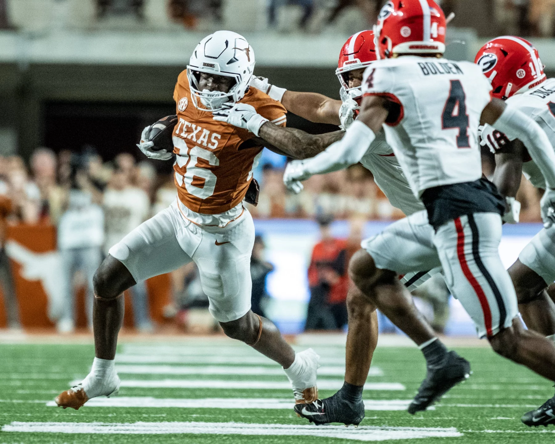 FootballR - NFL - Ein texanischer Footballspieler in Weiß und dunklem Orange versucht während des Spiels, einen Georgia Bulldog in Rot, Schwarz und Weiß mit ausgestrecktem Arm zu überwältigen. Auf dem Rasenfeld, umgeben von verschwommenen Zuschauern, hält der Austin-Athlet den Football in seinem rechten Arm und drängt sich durch die gegnerischen Spieler. Diese Beschreibung wurde automatisch generiert.