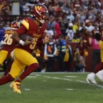 FootballR - NFL Washington Commanders - Quarterback Jayden Daniels saust in rot-gelber Uniform zielstrebig über das grüne Feld. Umgeben von blau gekleideten Gegnern und Teamkollegen, die ihm entscheidende Blockunterstützung geben, will er die Panthers zum Sieg führen. Eine verschwommene Menge jubelnder Zuschauer umringt das Geschehen. Diese Beschreibung wurde automatisch generiert.