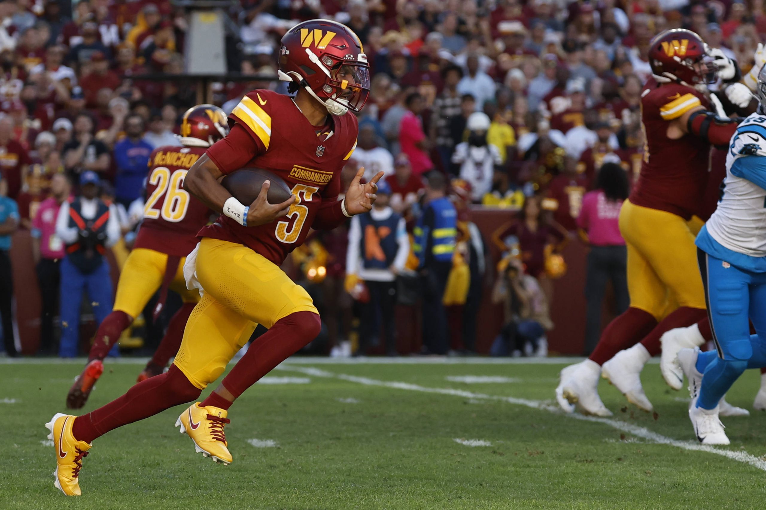 FootballR - NFL Washington Commanders - Quarterback Jayden Daniels saust in rot-gelber Uniform zielstrebig über das grüne Feld. Umgeben von blau gekleideten Gegnern und Teamkollegen, die ihm entscheidende Blockunterstützung geben, will er die Panthers zum Sieg führen. Eine verschwommene Menge jubelnder Zuschauer umringt das Geschehen. Diese Beschreibung wurde automatisch generiert.