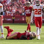 Während eines Spiels stehen Footballspieler gegnerischer Mannschaften auf dem Feld. Brandon Aiyuk liegt in seiner rot-goldenen Uniform auf dem Boden, während vier Spieler in rot-weißen Uniformen um ihn herum stehen und in verschiedene Richtungen blicken. Im Hintergrund ist die Menge im Stadion zu sehen. Diese Beschreibung wurde automatisch generiert.