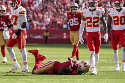 Während eines Spiels stehen Footballspieler gegnerischer Mannschaften auf dem Feld. Brandon Aiyuk liegt in seiner rot-goldenen Uniform auf dem Boden, während vier Spieler in rot-weißen Uniformen um ihn herum stehen und in verschiedene Richtungen blicken. Im Hintergrund ist die Menge im Stadion zu sehen. Diese Beschreibung wurde automatisch generiert.