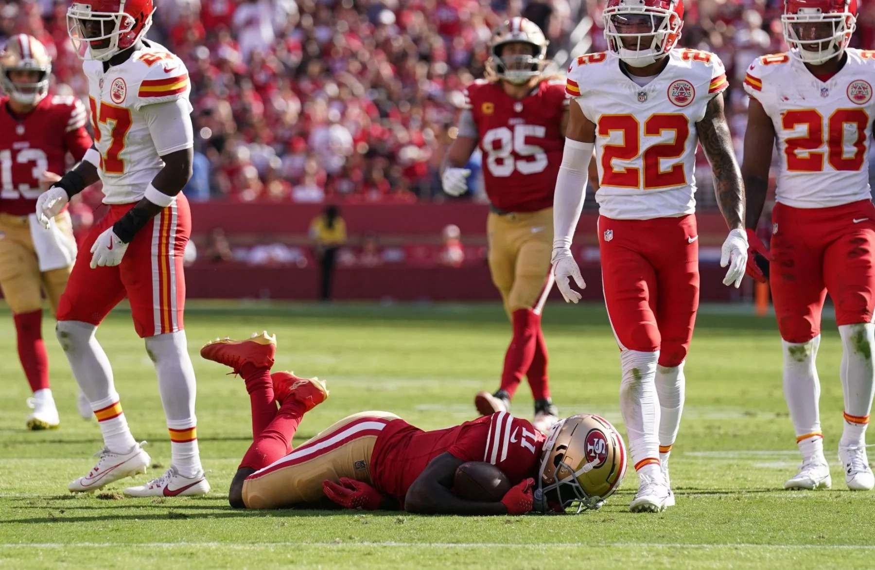 Während eines Spiels stehen Footballspieler gegnerischer Mannschaften auf dem Feld. Brandon Aiyuk liegt in seiner rot-goldenen Uniform auf dem Boden, während vier Spieler in rot-weißen Uniformen um ihn herum stehen und in verschiedene Richtungen blicken. Im Hintergrund ist die Menge im Stadion zu sehen. Diese Beschreibung wurde automatisch generiert.