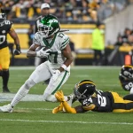 FootballR - NFL - Davante Adams rennt in einer weiß-grünen Uniform mit dem Ball über das Feld, während ein Gegenspieler in Schwarz-Gelb auf dem Rasen zu einem Tackling ansetzt. Im Stadion herrscht reges Treiben, die Teamfarben sind leuchtend und die Fans verfolgen gespannt jede Bewegung. Diese Beschreibung wurde automatisch generiert.