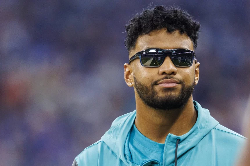 Free Agency - Tua Tagovailoa mit kurzen lockigen Haaren und Bart trägt eine dunkle Sonnenbrille und ein blaugrünes Sportjackett in den Farben der Dolphins. Er hat einen neutralen Gesichtsausdruck und steht vor einem verschwommenen Hintergrund, möglicherweise einem Stadion oder einer Sportstätte, die an den Ort erinnert, an dem Quarterback Tua Tagovailoa spielen könnte. Diese Beschreibung wurde mit der FootballR KI automatisch generiert.