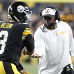 FootballR - NFL - Ein Spieler der Pittsburgh Steelers mit der Trikotnummer 3 namens Russell Wilson erhält während der NFL Woche 7 einHigh Five von Mike Tomlin. Der Trainer, in einem weißen Steelers-Shirt und einer schwarzen Kappe, wirkt konzentriert und hält die Karte mit seiner rechten Hand auf dem Footballfeld, mit verschwommenen Zuschauern im Hintergrund. Diese Beschreibung wurde automatisch generiert.