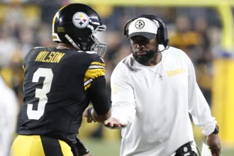 FootballR - NFL - Ein Spieler der Pittsburgh Steelers mit der Trikotnummer 3 namens Russell Wilson erhält während der NFL Woche 7 einHigh Five von Mike Tomlin. Der Trainer, in einem weißen Steelers-Shirt und einer schwarzen Kappe, wirkt konzentriert und hält die Karte mit seiner rechten Hand auf dem Footballfeld, mit verschwommenen Zuschauern im Hintergrund. Diese Beschreibung wurde automatisch generiert.