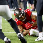 FootballR - NFL - Mike Evans liegt im roten Trikot auf dem Boden und hält den Ball fest. Zwei Spieler in dunklen Uniformen sind zu sehen, einer von links und ein anderer von rechts. Die Action spielt sich auf einem Rasenfeld während eines intensiven Nachtspiels ab. Diese Beschreibung wurde automatisch generiert.