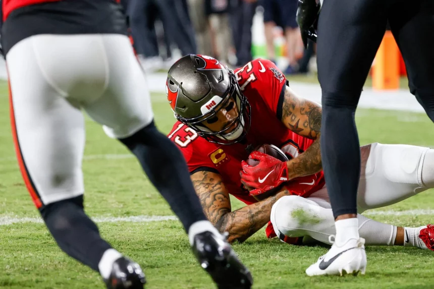 FootballR - NFL - Mike Evans liegt im roten Trikot auf dem Boden und hält den Ball fest. Zwei Spieler in dunklen Uniformen sind zu sehen, einer von links und ein anderer von rechts. Die Action spielt sich auf einem Rasenfeld während eines intensiven Nachtspiels ab. Diese Beschreibung wurde automatisch generiert.