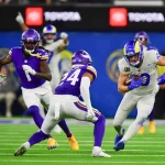 FootballR - NFL - Cooper Kupp in weißer Uniform und blauem Helm läuft mit dem Ball, während zwei Gegner in violetten Uniformen und Helmen ihn tackeln. Puka Nacua freut sich im Hintergrund über den gelungenen Spielzug. Das grüne Feld weist weiße Yard-Linien auf, und „TOYOTA“-Banner zieren den Hintergrund neben verschwommenen Zuschauern. Diese Beschreibung wurde automatisch generiert.