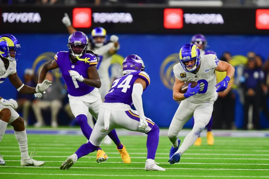 FootballR - NFL - Cooper Kupp in weißer Uniform und blauem Helm läuft mit dem Ball, während zwei Gegner in violetten Uniformen und Helmen ihn tackeln. Puka Nacua freut sich im Hintergrund über den gelungenen Spielzug. Das grüne Feld weist weiße Yard-Linien auf, und „TOYOTA“-Banner zieren den Hintergrund neben verschwommenen Zuschauern. Diese Beschreibung wurde automatisch generiert.