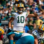 Ein Quarterback in grün-goldener Uniform, Nummer 10, dirigiert seine Teamkollegen auf dem Spielfeld. Jordan Love ist von Spielern beider Teams umgeben. Die Szene spielt sich in einem Stadion voller Zuschauer ab, das Sonnenlicht wirft Schatten auf das Spielfeld. Diese Beschreibung wurde mit der FootballR KI automatisch generiert.