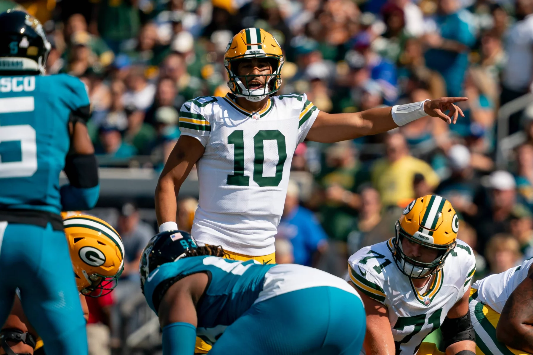 Ein Quarterback in grün-goldener Uniform, Nummer 10, dirigiert seine Teamkollegen auf dem Spielfeld. Jordan Love ist von Spielern beider Teams umgeben. Die Szene spielt sich in einem Stadion voller Zuschauer ab, das Sonnenlicht wirft Schatten auf das Spielfeld. Diese Beschreibung wurde mit der FootballR KI automatisch generiert.