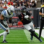 Stefon Diggs in schwarzer Uniform mit roten Akzenten, die an Stefon Diggs' legendären Stil erinnert, läuft mit dem Ball, dicht gefolgt von einem Spieler in Weiß und Blau. Das Geschehen findet auf einem grünen Feld mit weißen Spielfeldmarkierungen statt, während ein Schiedsrichter zusieht und Zuschauer den Hintergrund füllen. Diese Beschreibung wurde mit der FootballR KI automatisch generiert.