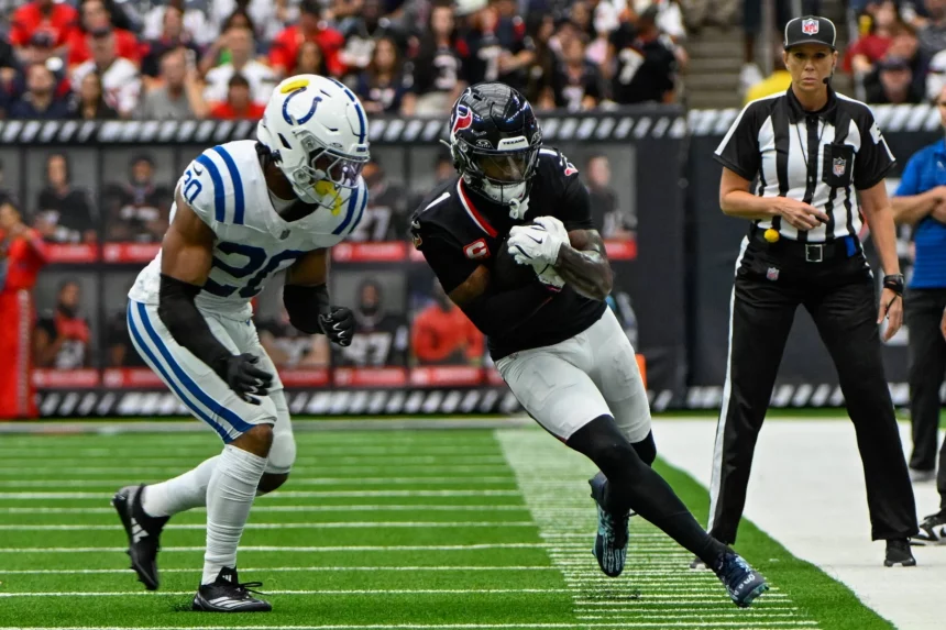 Stefon Diggs in schwarzer Uniform mit roten Akzenten, die an Stefon Diggs' legendären Stil erinnert, läuft mit dem Ball, dicht gefolgt von einem Spieler in Weiß und Blau. Das Geschehen findet auf einem grünen Feld mit weißen Spielfeldmarkierungen statt, während ein Schiedsrichter zusieht und Zuschauer den Hintergrund füllen. Diese Beschreibung wurde mit der FootballR KI automatisch generiert.