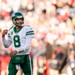 Aaron Rodgers in einer grün-weißen Uniform der New York Jets mit der Nummer 8 steht auf dem Feld. Er hält den Kinnriemen seines Helms und blickt zur Seite. Die Menge im Hintergrund ist verschwommen, was seine konzentrierte Präsenz und Hingabe für die Jets unterstreicht. Jets-Colts Game verschoben. Diese Beschreibung wurde mit der FootballR KI automatisch generiert.