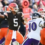 New York Giants - Ein Footballspieler in einem schwarzen Trikot mit der Nummer 5 , Jameis Winston, als er sich während eines Spiels auf einen Pass vorbereitet. Er wird von einem Abwehrspieler in einem weißen Trikot mit der Nummer 10 verfolgt. Spieler in orange-weißen Uniformen, wie die der Cleveland Browns, sind ebenfalls zu sehen, wobei die Feld- und Downmarkierung zu sehen ist. Diese Beschreibung wurde mit der FootballR KI automatisch generiert.