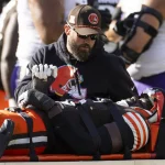 Jeremiah Owusu-Koramoah, ein Footballspieler in Schwarz, liegt auf einer orangefarbenen Bahre. Ein Mann in schwarzer Jacke und Mütze kniet neben ihm und hält ihm die Hand. Im Hintergrund sind andere Spieler in weißen und violetten Uniformen zu sehen, die sich besorgt entwickeln. Diese Beschreibung wurde mit der FootballR KI automatisch generiert.