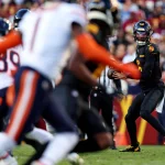 In Woche 8 stand Jayden Daniels in schwarz-gelber Uniform bereit, den Ball während eines Spiels zu werfen. Gegner in weißen Uniformen mit orangefarbenen Streifen nähern sich von links. Im Stadion wimmelt es von Zuschauern, und das Spielfeld ist von einer rot-gelben Seitenlinie begrenzt. Diese Beschreibung wurde mit der FootballR KI automatisch generiert.