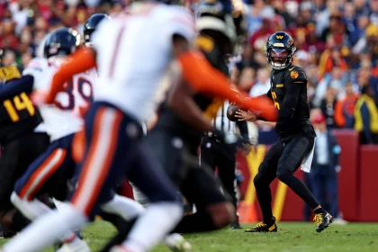 In Woche 8 stand Jayden Daniels in schwarz-gelber Uniform bereit, den Ball während eines Spiels zu werfen. Gegner in weißen Uniformen mit orangefarbenen Streifen nähern sich von links. Im Stadion wimmelt es von Zuschauern, und das Spielfeld ist von einer rot-gelben Seitenlinie begrenzt. Diese Beschreibung wurde mit der FootballR KI automatisch generiert.