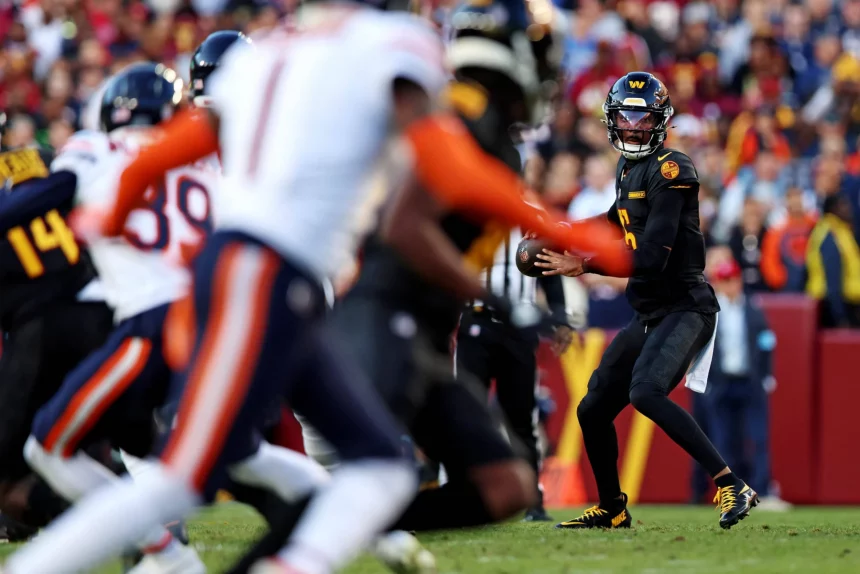 In Woche 8 stand Jayden Daniels in schwarz-gelber Uniform bereit, den Ball während eines Spiels zu werfen. Gegner in weißen Uniformen mit orangefarbenen Streifen nähern sich von links. Im Stadion wimmelt es von Zuschauern, und das Spielfeld ist von einer rot-gelben Seitenlinie begrenzt. Diese Beschreibung wurde mit der FootballR KI automatisch generiert.