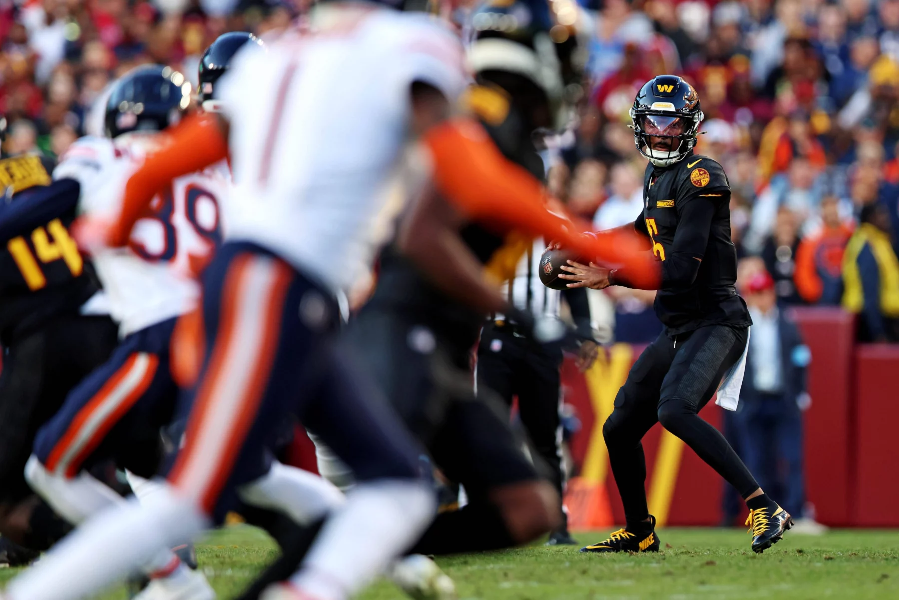 In Woche 8 stand Jayden Daniels in schwarz-gelber Uniform bereit, den Ball während eines Spiels zu werfen. Gegner in weißen Uniformen mit orangefarbenen Streifen nähern sich von links. Im Stadion wimmelt es von Zuschauern, und das Spielfeld ist von einer rot-gelben Seitenlinie begrenzt. Diese Beschreibung wurde mit der FootballR KI automatisch generiert.