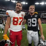 Zwei Footballspieler stehen am National Tight Ends Day zusammen auf dem Spielfeld. Travis Kelce trägt ein rot-weißes Trikot mit der Nummer 87, Brock Bowers ein schwarzes Trikot mit der Nummer 89. Sie lächeln herzlich und legen sich die Arme um die Schultern, während die Menge voller Vorfreude summt. Diese Beschreibung wurde mit der FootballR KI automatisch generiert.