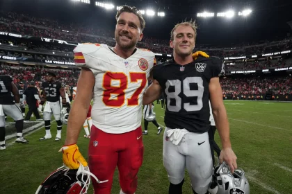 Zwei Footballspieler stehen am National Tight Ends Day zusammen auf dem Spielfeld. Travis Kelce trägt ein rot-weißes Trikot mit der Nummer 87, Brock Bowers ein schwarzes Trikot mit der Nummer 89. Sie lächeln herzlich und legen sich die Arme um die Schultern, während die Menge voller Vorfreude summt. Diese Beschreibung wurde mit der FootballR KI automatisch generiert.