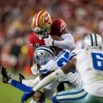 Der Footballspieler Deebo Samuel oder Jordan Mason in einer roten Uniform hält den Ball fest, während er nach vorne stürmt, verfolgt von zwei Spielern in weißen und blauen Uniformen auf dem Feld. Die Szene ist intensiv, ein Spieler streckt den Arm aus, um Mason zu Fall zu bringen. Der Hintergrund ist durch eifrige Zuschauer verschwommen. Diese Beschreibung wurde mit der FootballR KI automatisch generiert.