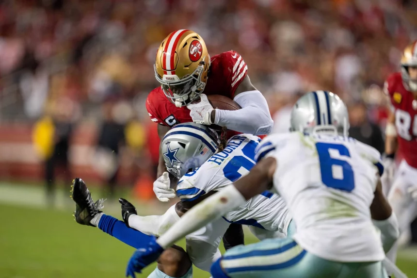 Der Footballspieler Deebo Samuel oder Jordan Mason in einer roten Uniform hält den Ball fest, während er nach vorne stürmt, verfolgt von zwei Spielern in weißen und blauen Uniformen auf dem Feld. Die Szene ist intensiv, ein Spieler streckt den Arm aus, um Mason zu Fall zu bringen. Der Hintergrund ist durch eifrige Zuschauer verschwommen. Diese Beschreibung wurde mit der FootballR KI automatisch generiert.