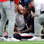 Ein Footballspieler in schwarzer Uniform, Stefon Diggs , sitzt auf dem Feld, umgeben von besorgten Trainern und medizinischem Personal, die seinen Zustand besprechen. Eine Gatorade-Flasche liegt während dieses intensiven Saison-Spiels gegen die Texans in der Nähe auf dem grünen Rasen des Sportstadions. Diese Beschreibung wurde mit der FootballR KI automatisch generiert.