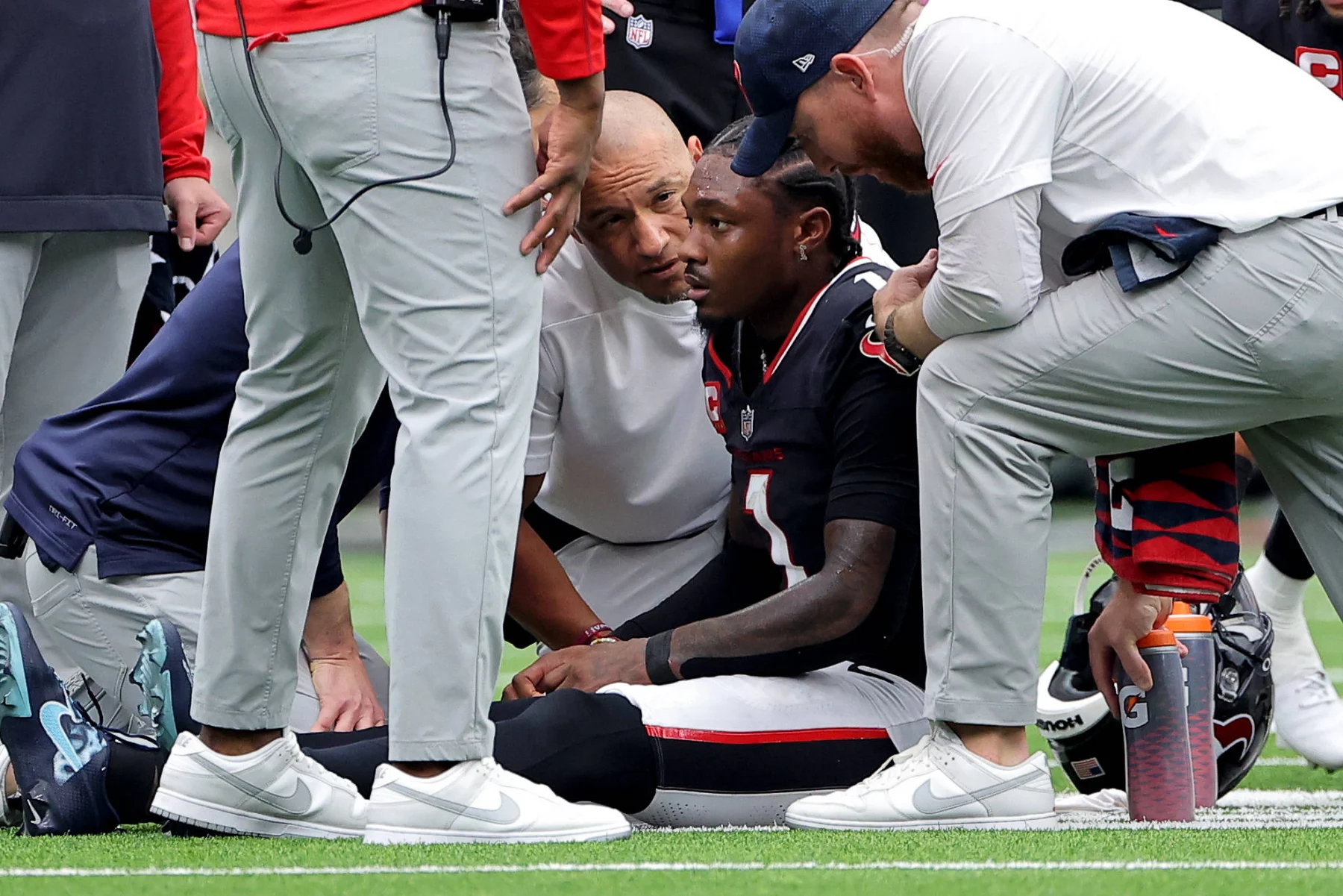 Ein Footballspieler in schwarzer Uniform, Stefon Diggs , sitzt auf dem Feld, umgeben von besorgten Trainern und medizinischem Personal, die seinen Zustand besprechen. Eine Gatorade-Flasche liegt während dieses intensiven Saison-Spiels gegen die Texans in der Nähe auf dem grünen Rasen des Sportstadions. Diese Beschreibung wurde mit der FootballR KI automatisch generiert.